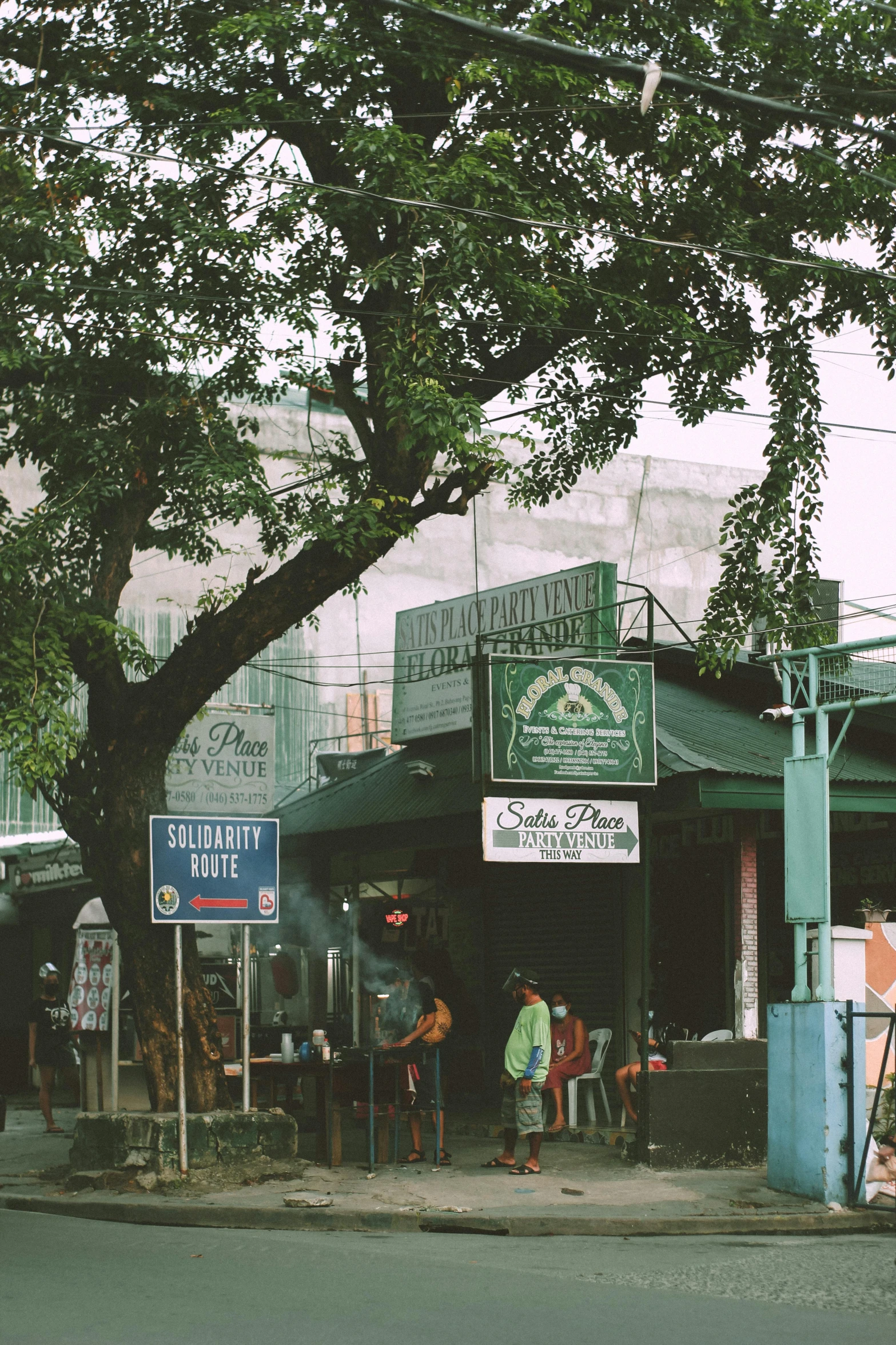 a group of people standing on the side of a road, green alleys, lots of signs and shops, big green tree, puerto rico