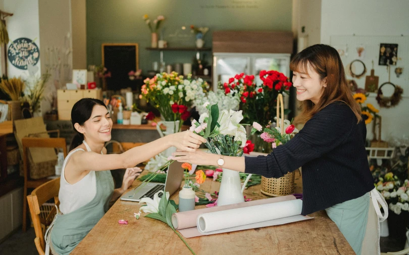 two women sitting at a table in a flower shop, pexels contest winner, reaching out to each other, simple stylized, staff, over the shoulder