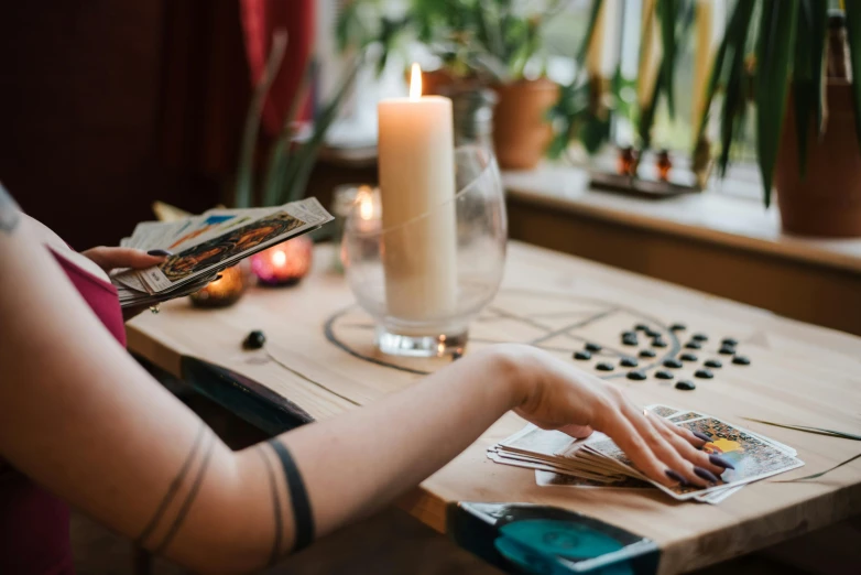 a woman sitting at a table with cards and a candle, trending on pexels, witchy clothing, photograph of a sleeve tattoo, on a wooden tray, pair of keycards on table