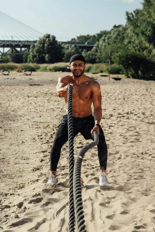 a man holding a rope on top of a sandy beach, fit physique, instagram story, working out in the field, playful smirk