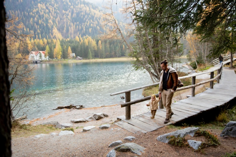 a man walking a dog across a wooden bridge, a picture, by Carlo Martini, lago di sorapis, fall season, a cozy, tan