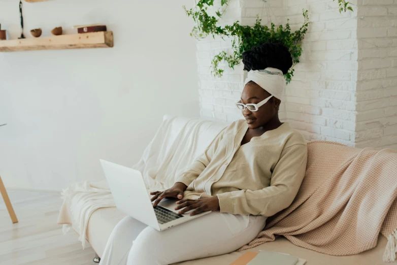 a woman sitting on a couch using a laptop, by Carey Morris, trending on pexels, afrofuturism, wearing white pajamas, brown and white color scheme, girl with glasses, developers