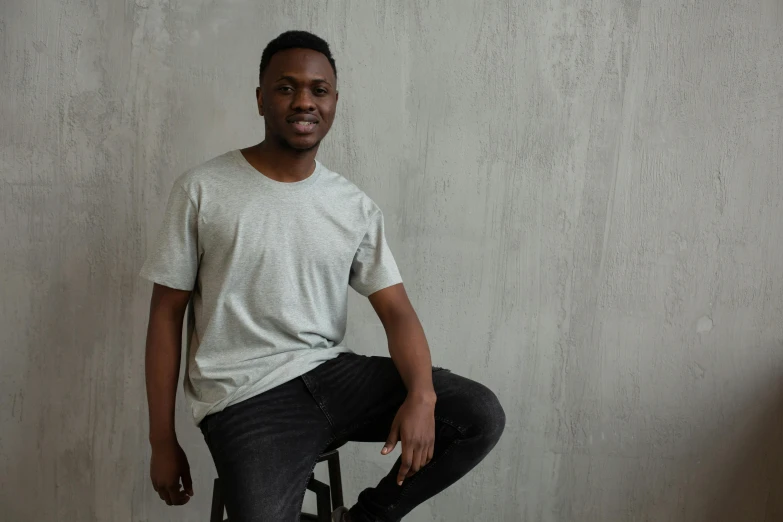 a man sitting on a stool in front of a wall, jeans and t shirt, in gunmetal grey, emmanuel shiru, medium head to shoulder shot