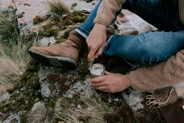 a person sitting on a rock with a can of beer, trending on pexels, land art, picking up a can beans, leather clothing and boots, avatar image, soup