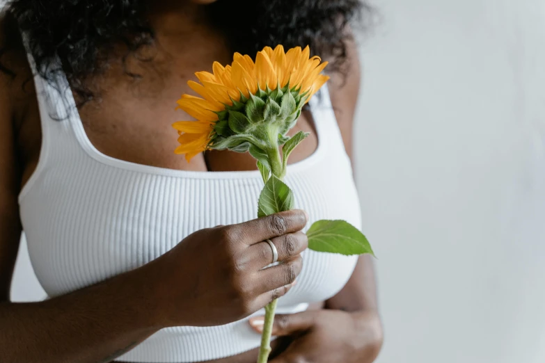 a close up of a person holding a flower, trending on pexels, bare midriff, young black woman, helianthus flowers, white and orange breastplate