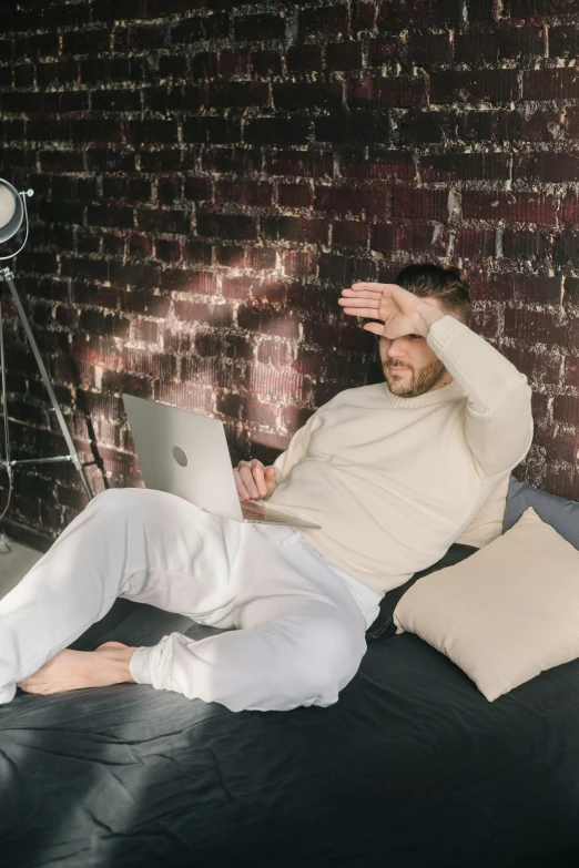 a man sitting on top of a bed with a laptop, doing a sassy pose, wearing white pajamas, waiting behind a wall, trending photo