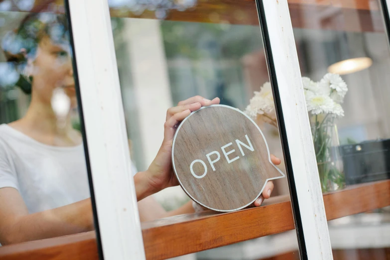 a woman holding an open sign in front of a window, trending on unsplash, award winning shopfront design, on a wooden plate, portal opening, thumbnail