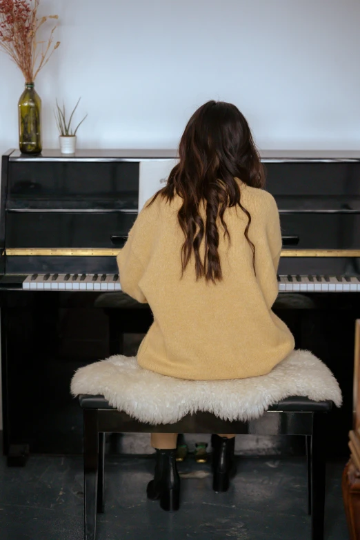 a woman sitting at a piano in a living room, by Alice Mason, trending on pexels, wavy hair yellow theme, back pose, cardigan, brunette