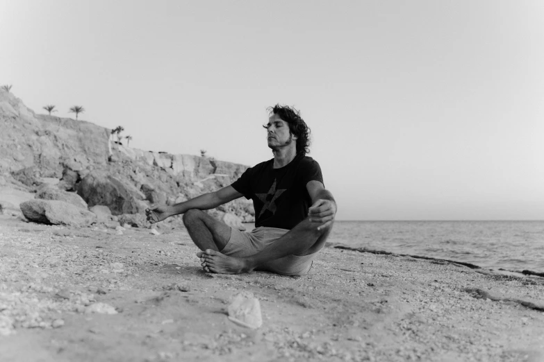 a black and white photo of a man sitting on the beach, figuration libre, zack de la rocha, sitting cross-legged, red sea, profile picture