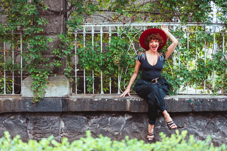 a woman in a red hat sitting on a stone wall, by Lucia Peka, pexels contest winner, art nouveau, dark short curly hair smiling, in chippendale sydney, black hat, silly
