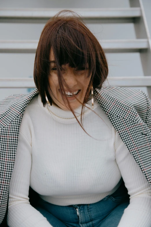 a woman sitting on the steps of a building, inspired by Zoë Mozert, trending on pexels, happening, head bent back in laughter, wearing a turtleneck and jacket, with dark brown bangs, wearing a white button up shirt