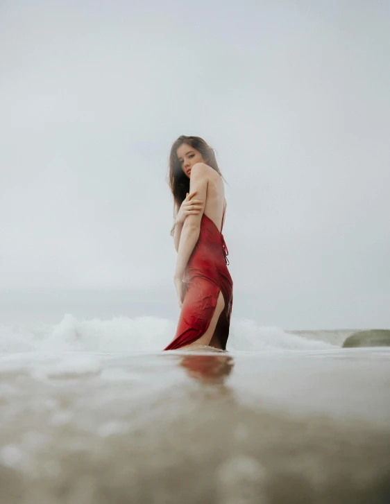 a woman in a red dress standing in the ocean, by Robbie Trevino, low quality photo, gif, ombre velvet gown, on a cloudy day