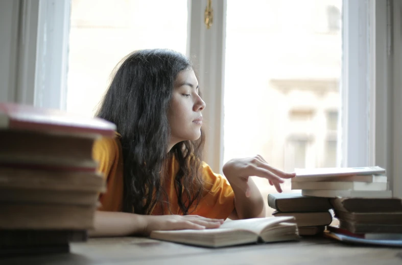 a woman sitting at a table reading a book, pexels contest winner, academic art, holding a stack of books, leaning against the window, teenage girl, pictured from the shoulders up