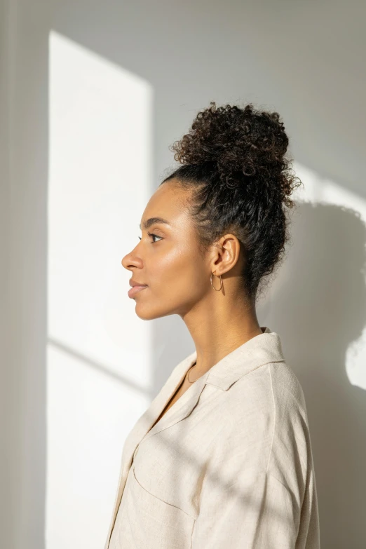 a woman standing in front of a white wall, hair tied in a bun, detailed product image, diffuse natural sun lights, (dark shorter curly hair)