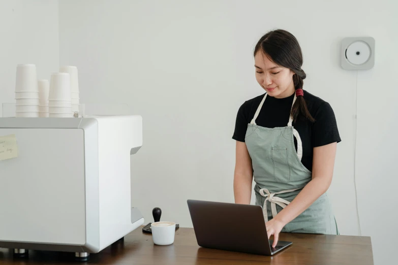 a woman in an apron working on a laptop, pexels contest winner, aussie baristas, standing on a desk, avatar image, half asian