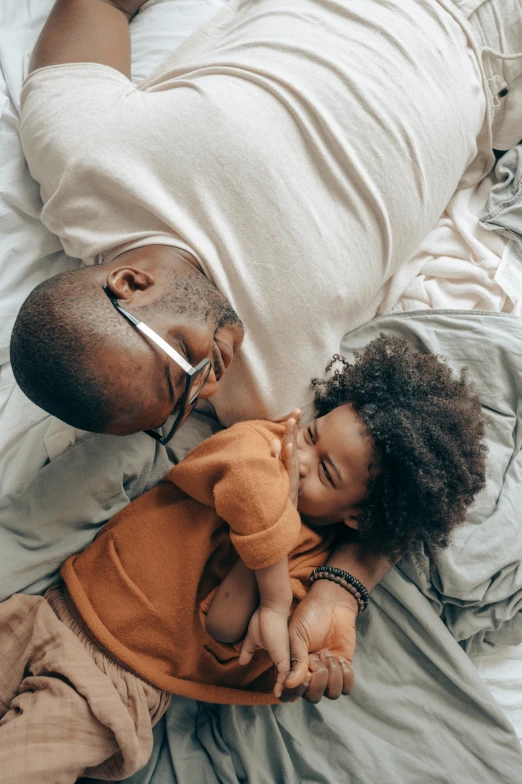 a man laying on top of a bed holding a baby, pexels contest winner, visual art, light skinned african young girl, flat lay, kissing together, father with child