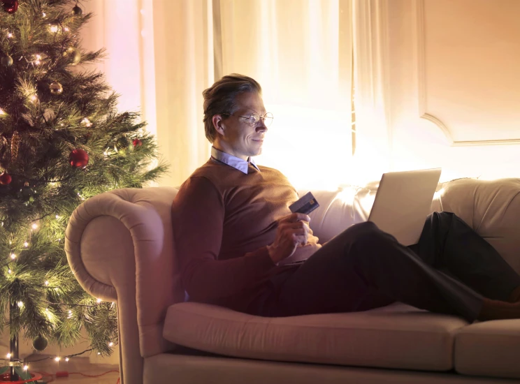 a man sitting on a couch in front of a christmas tree, technology, warmly lit posh study, tv still, skies behind