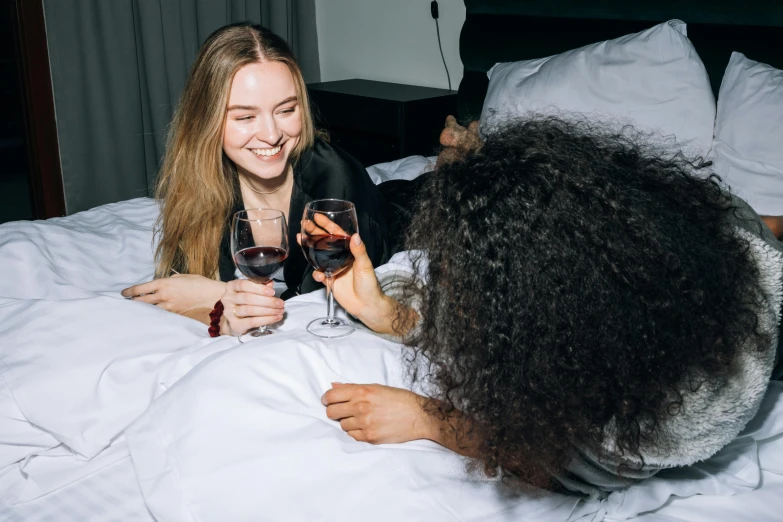 a couple of women sitting on top of a bed next to each other, by Cosmo Alexander, pexels contest winner, happening, holding a glass of red wine, sydney sweeney, wavy hair spread out, with black