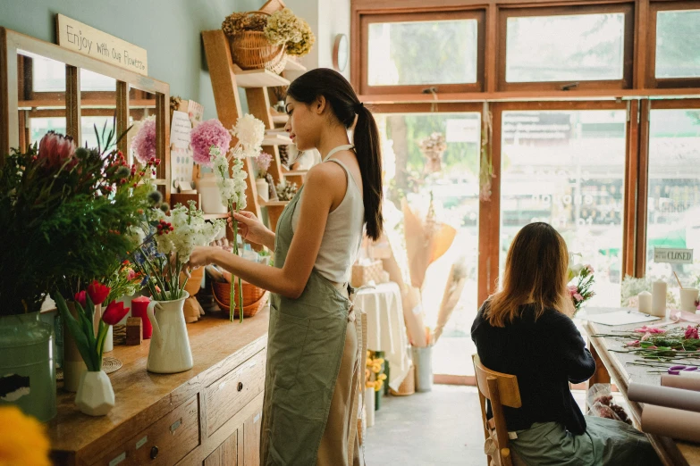 a woman standing in front of a table filled with flowers, trending on unsplash, shop front, small manufacture, facing left, customers