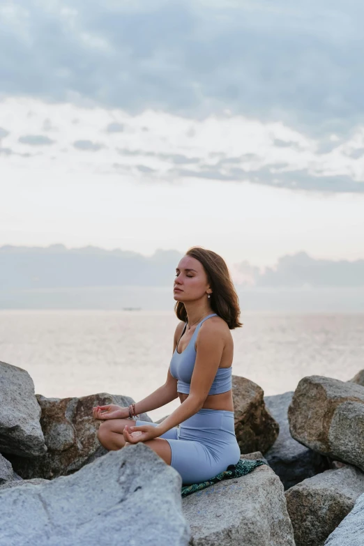 a woman is sitting on some rocks by the water, unsplash, renaissance, padmasana, avatar image, grey, 8k octan photo