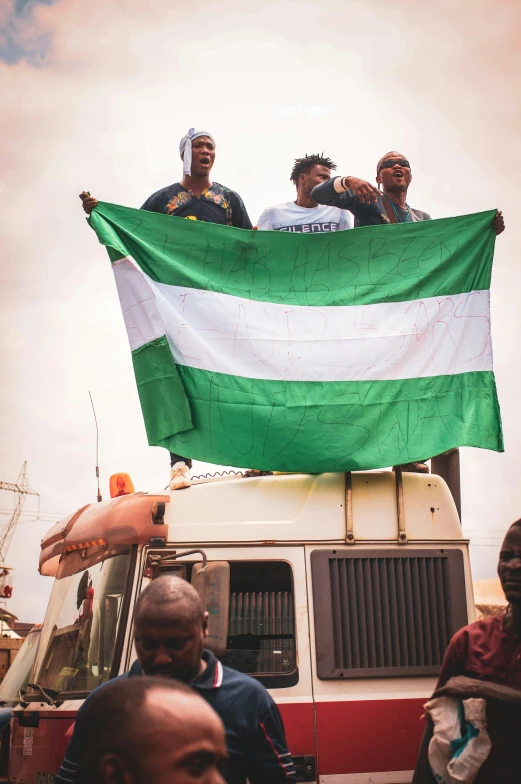 a group of people sitting on top of a truck, by Matija Jama, trending on unsplash, green flag, african, holding a white flag, he is a long boi ”