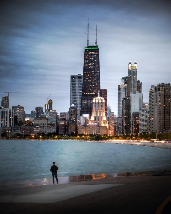 a man standing on a beach next to a body of water, by Greg Rutkowski, pexels contest winner, chicago skyline, lgbt, big buildings, late evening