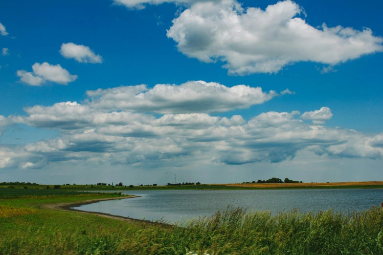 a large body of water sitting next to a lush green field, by Matthias Stom, unsplash, land art, cotton clouds, helmond, big sky, bartlomiej gawel