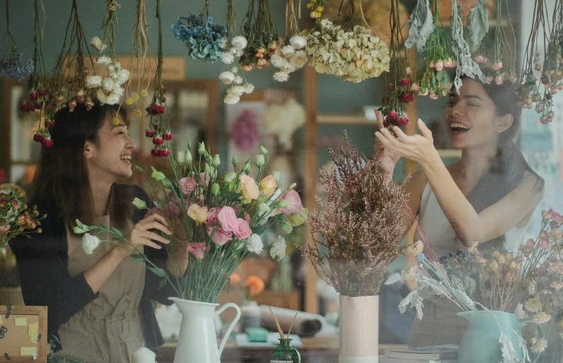 a couple of women sitting at a table with flowers, trending on pexels, things hanging from ceiling, happy friend, flower shop scene, dried flowers