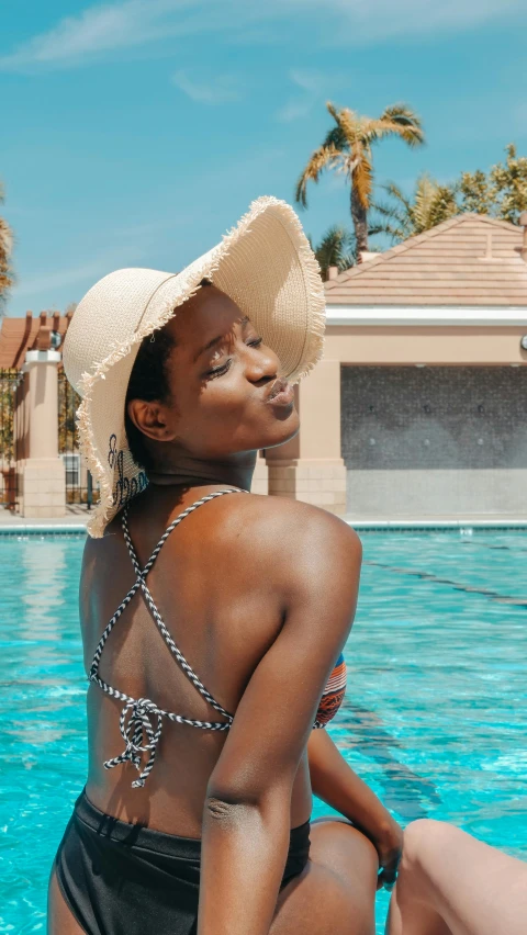 a woman sitting on the edge of a swimming pool, by Lily Delissa Joseph, pexels contest winner, afrofuturism, wearing a chocker and cute hat, back pose, thumbnail, brown skinned