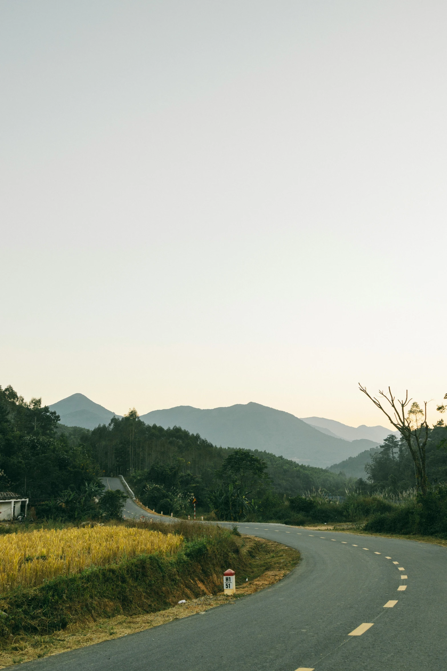 a man riding a skateboard down the middle of a road, a picture, unsplash, minimalism, distant town in valley and hills, vietnam, at sunrise in springtime, wide film still