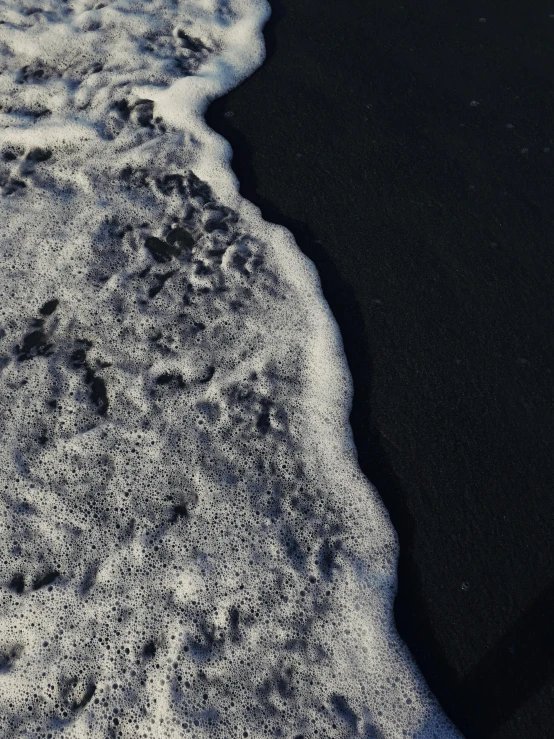 a person standing on a beach next to the ocean, inspired by Vija Celmins, pexels contest winner, land art, frosted texture, drone photo, black sand, surface with beer-texture