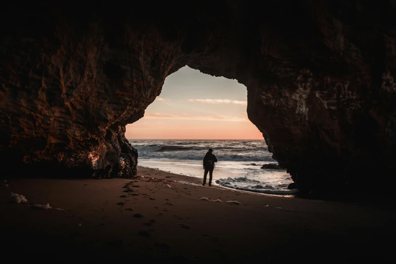 a person standing in front of a cave on a beach, by Daniel Seghers, pexels contest winner, hollister ranch, end of the day, abel tasman, portal opening