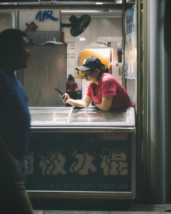 a woman looking at her cell phone while standing in front of a food stand, inspired by Liam Wong, trending on unsplash, queer woman, fish market, in a kitchen, freezing