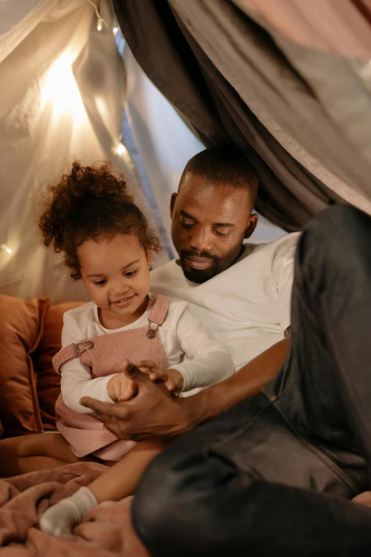 a man and a little girl sitting on a couch, in a colorful tent, dark-skinned, dad energy, connectivity