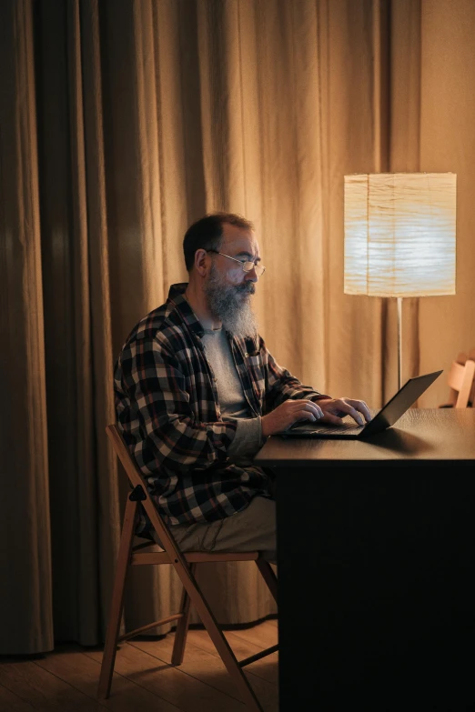 a man sitting at a desk using a laptop computer, by James Morris, light beard, multiple lights, lachlan bailey, sitting across the room
