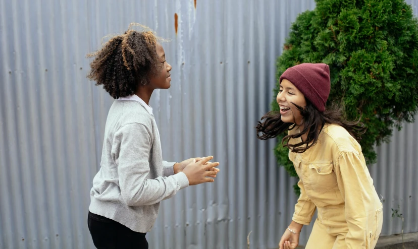 a couple of women standing next to each other on a street, by Alice Mason, pexels contest winner, interactive art, kids playing, beanie, earing a shirt laughing, vine twist