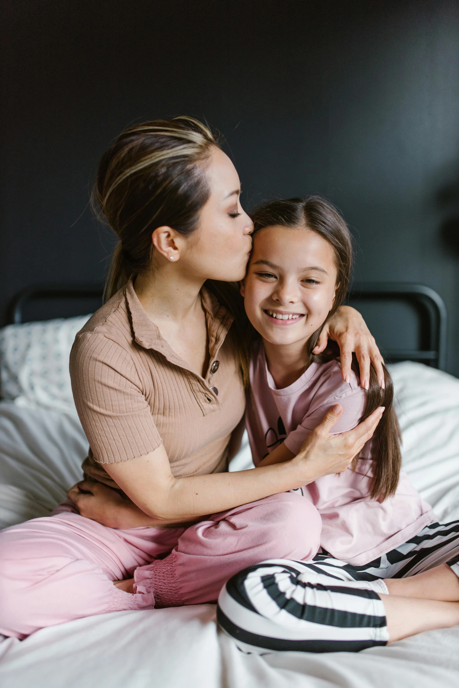 a woman sitting on top of a bed next to a little girl, kissing each other, gen z, arm around her neck, multiple stories