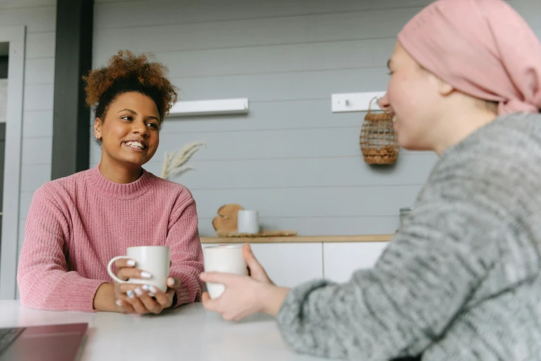 two women sitting at a table having a conversation, trending on pexels, pink and grey muted colors, with a white mug, welcoming smile, wearing a pink head band