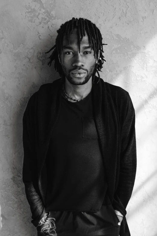 a man with dreadlocks standing in front of a wall, with a beard and a black shirt, lil uzi vert, black - and - white photograph, plain background