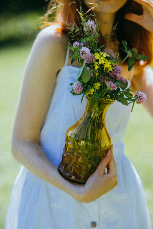 a woman in a white dress holding a vase of flowers, inspired by Elsa Beskow, unsplash, redhead woman, yellow, clover, old color photograph