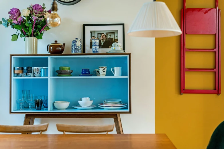 a table and chairs in a room with a clock on the wall, inspired by Lewis Henry Meakin, unsplash, maximalism, yellow and blue and cyan, shelf, 7 0's kitchen, complementary lighting