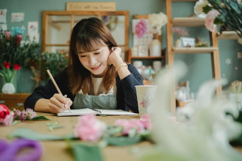 a woman sitting at a table writing on a piece of paper, inspired by Kim Jeong-hui, pexels contest winner, flower shop scene, looking happy, background image, student