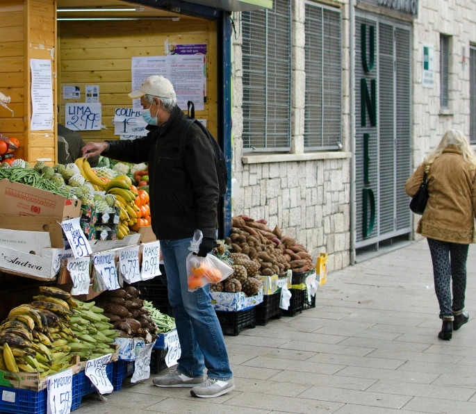 a man standing in front of a fruit and vegetable stand, a photo, by Niko Henrichon, unsplash, sukkot, square, people are wearing masks, people walking down a street