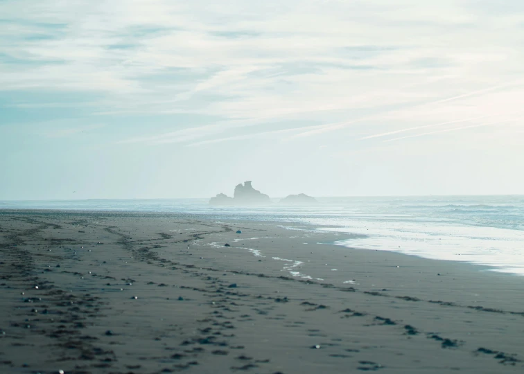 a man standing on top of a sandy beach next to the ocean, a picture, unsplash contest winner, minimalism, photo of shiprock, sparsely populated, people walking in the distance, foggy photo 8 k