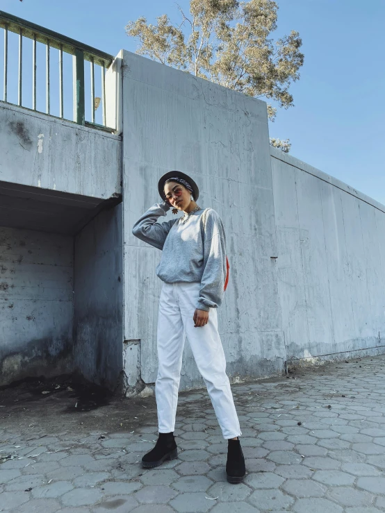 a woman standing in front of a wall talking on a cell phone, by Robbie Trevino, white pants, gray hoodie, instagram picture, uniform off - white sky