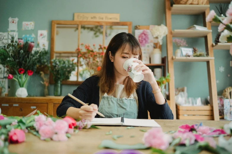 a woman sitting at a table with a cup of coffee, inspired by Kim Jeong-hui, pexels contest winner, flower shop scene, crafts and souvenirs, sketching, portrait image