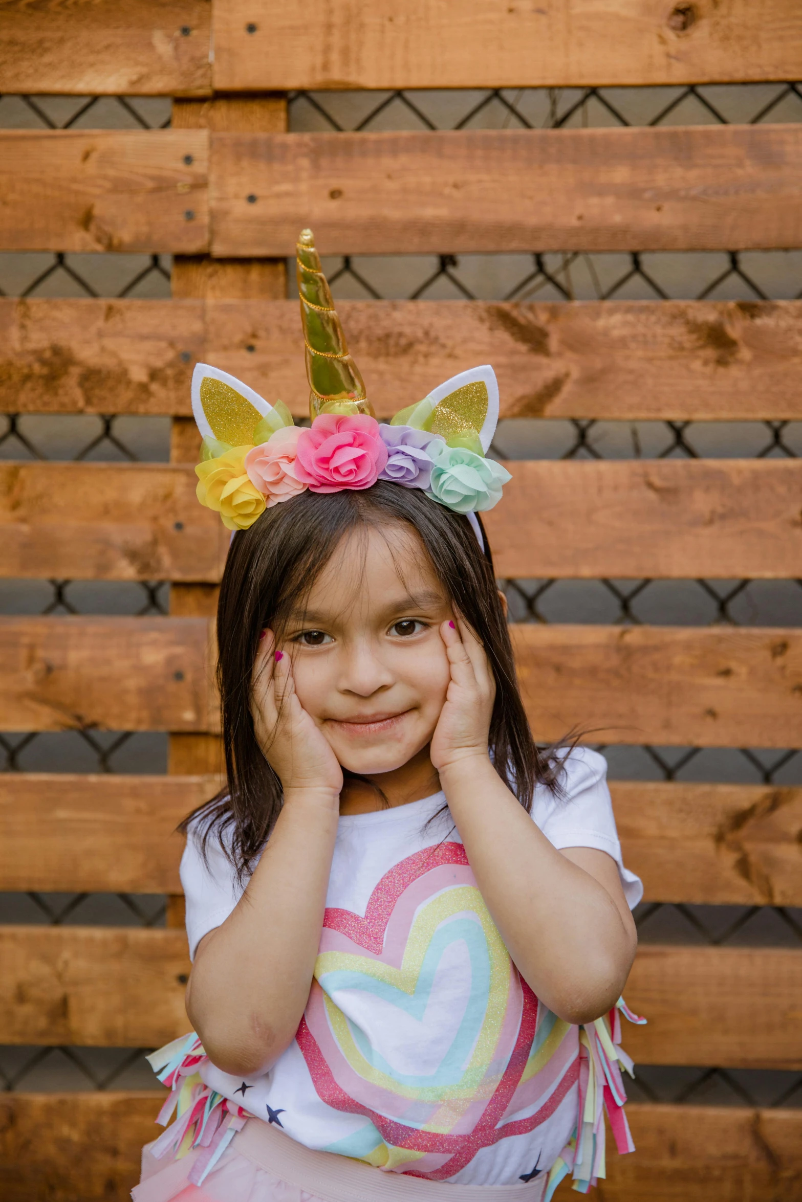 a little girl standing in front of a wooden fence, unicorn horn, wearing a paper crown, ready to model, ultra - quality