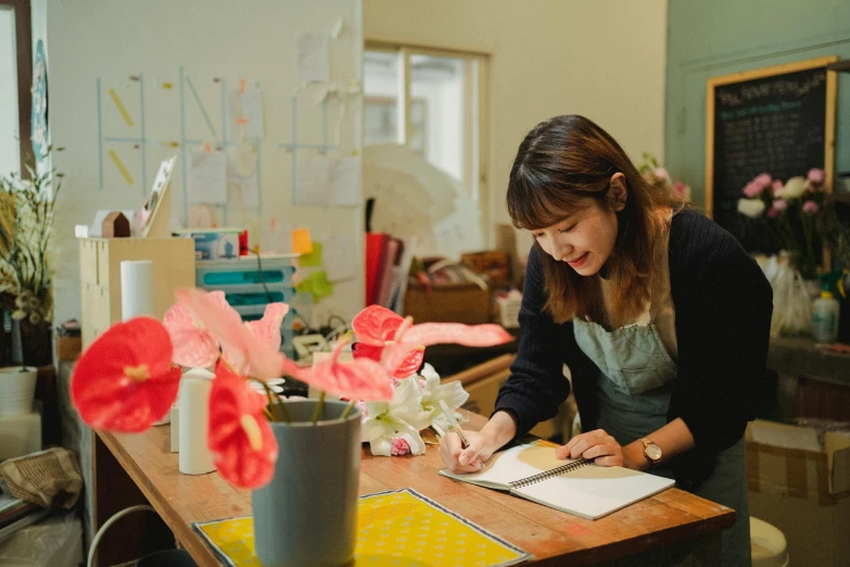 a woman sitting at a table writing on a piece of paper, a silk screen, inspired by Masamitsu Ōta, pexels contest winner, paper origami flowers, studio kyoto, looking happy, floral design