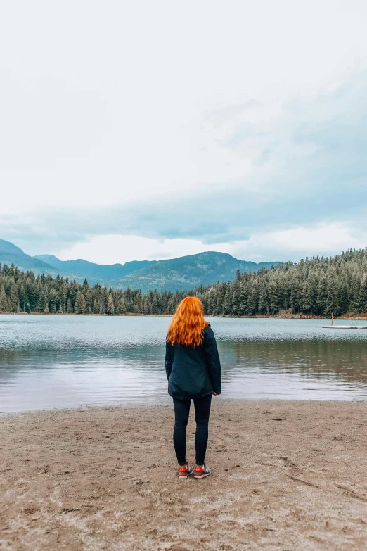 a woman standing on a beach next to a body of water, a picture, trending on unsplash, mountain lakes, ginger hair, erin moriarty, distant full body view