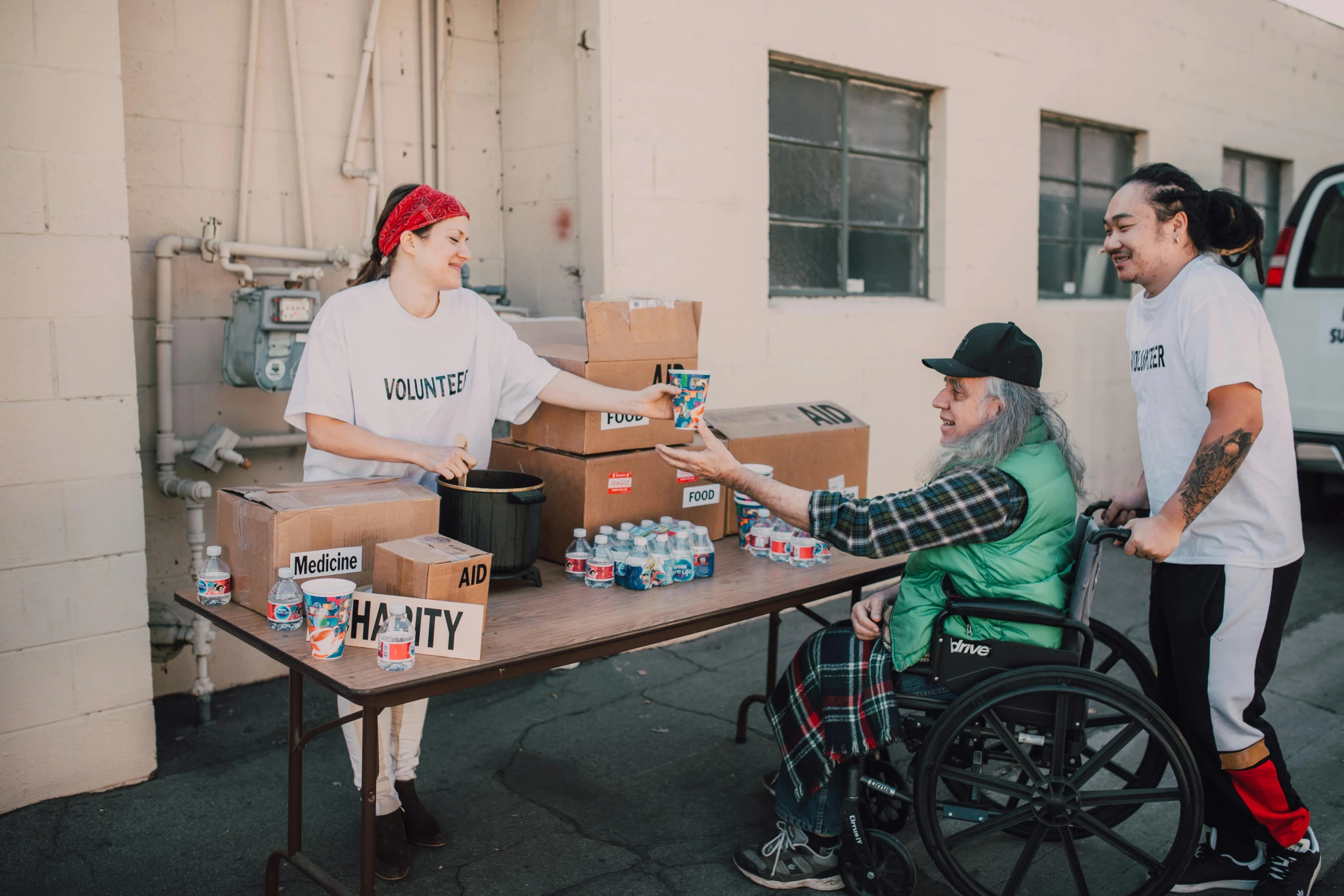 a group of people standing around a table with boxes on it, by Francis Helps, pexels contest winner, sitting in a wheelchair, california;, hunger, medium shot of two characters